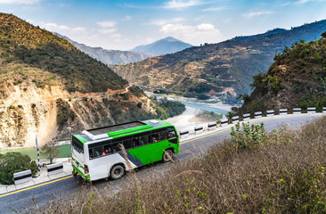 A white-green bus moves along the serpentine mountain road against the background of a river valley among the mountains.