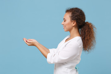 Wall Mural - Side view of african american doctor woman isolated on blue background. Female doctor in white medical gown hold something in palms. Healthcare personnel health medicine concept. Mock up copy space.