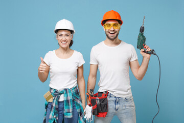 Funny young couple woman man in protective helmet hardhat hold electric drill isolated on blue background. Instruments accessories for renovation apartment room. Repair home concept. Showing thumb up.