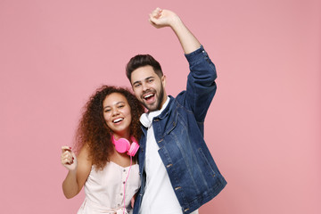 Wall Mural - Laughing young couple two friends european guy african american girl in casual clothes with headphones posing isolated on pastel pink background studio. People lifestyle concept. Rising hands dancing.