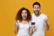 © ViDi Studio - Excited young couple two friends european guy african american girl in white t-shirts posing isolated on yellow background studio. People lifestyle concept. Hold credit bank card doing winner gesture.