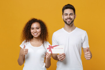 Wall Mural - Smiling young couple two friends european guy african american girl in white t-shirts posing isolated on yellow background studio. People lifestyle concept. Hold gift certificate, showing thumbs up.