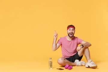 Excited fitness guy sportsman in headband t-shirt in home gym isolated on yellow background. Workout sport motivation concept. Sit with smart watch, water mobile phone dumbbells point index finger up.