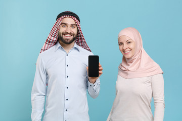 Wall Mural - Smiling couple friends arabian muslim man wonam in keffiyeh kafiya ring igal agal hijab clothes isolated on blue background. People religious lifestyle concept. Hold mobile phone with empty screen.