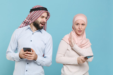 Canvas Print - Shocked concerned couple friends arabian muslim man wonam in keffiyeh kafiya ring igal agal hijab clothes isolated on blue background. People religious concept. Using mobile phone, typing sms message.