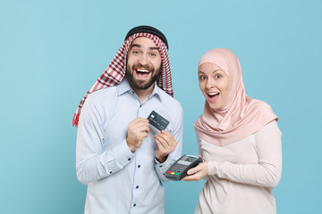 Wall Mural - Excited couple friends arabian muslim man wonam in keffiyeh kafiya ring igal hijab clothes isolated on blue background. People religious concept. Hold payment terminal to process credit card payments.