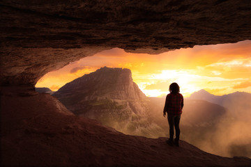 Wall Mural - Adventurous Girl Looking out at a dramatic American Mountain Landscape from a rocky cave during a colorful sunset. Fantasy Adventure Composite.