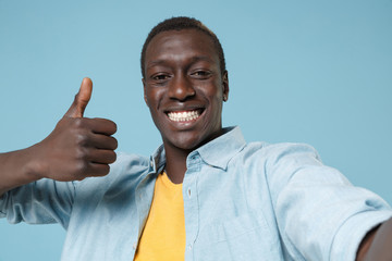 Close up of smiling young african american man guy in casual shirt, yellow t-shirt posing isolated on blue background. People lifestyle concept. Doing selfie shot on mobile phone, showing thumb up.