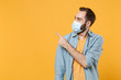 © ViDi Studio - Young man in sterile face mask posing isolated on yellow background studio portrait. Epidemic pandemic spreading coronavirus 2019-ncov sars covid-19 flu virus concept. Pointing index finger aside up.