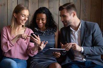 Happy multiracial young people sit on couch have fun using electronic gadgets together, smiling diverse multiethnic friends relax in cafe laugh joke showing pictures on devices, technology concept