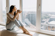 © Bostan Natalia - Young woman sitting on a windowsill by the window with a city view, drinking coffee or tea in the morning.