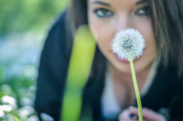 Close up portrait of a young woman with a dandelion laying on the grass. Positive girl having fun outside in a sunny day. Focus on the flower.