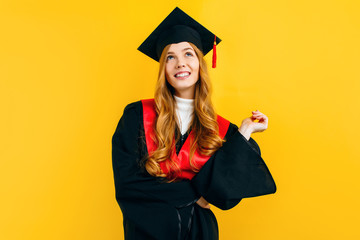 Wall Mural - A thoughtful, graduate in a master's dress, on a yellow background. Concept of the graduation ceremony