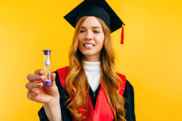 Wall Mural - A happy graduate holds an hourglass in her hand on an isolated yellow background