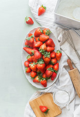 Fresh organic garden strawberries in white plate on light kitchen table with baking tools and ingredients. Top view. Healthy seasonal food.