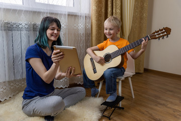 cute boy plays the guitar, mom holds a tablet and shows to his son and smiles. a fun online lesson o