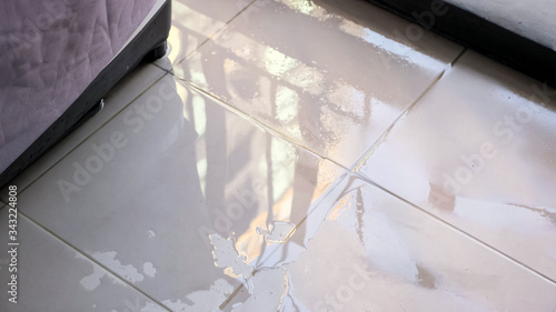 trembling water puddle after pipe burst covers white tile near furniture in room reflecting window and blue sky close view