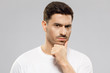 © Damir Khabirov - Close-up of young man in white t-shirt, holding his chin, looking at camera with expression of suspicion, mistrusting and doubting, isolated on gray background