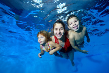 Sports family: a smiling mother and her two young children swim underwater in the pool. They hug, smile and pose for the camera on a blue background. Close up. Digital photo.