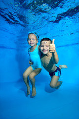 A little boy and his younger sister pose underwater in a children's pool. The boy smiles broadly, looks at the camera, and gives a thumbs-up. Closeup. Digital photo.