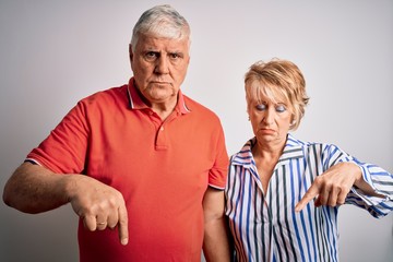 Wall Mural - Senior beautiful couple standing together over isolated white background Pointing down looking sad and upset, indicating direction with fingers, unhappy and depressed.