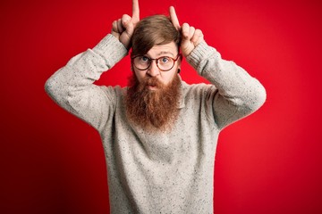 Poster - Handsome Irish redhead man with beard wearing casual sweater and glasses over red background doing funny gesture with finger over head as bull horns