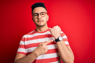 Young handsome man wearing casual striped t-shirt and glasses over isolated red background In hurry pointing to watch time, impatience, looking at the camera with relaxed expression