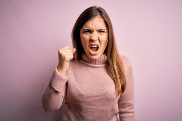 Sticker - Beautiful young woman wearing turtleneck sweater over pink isolated background angry and mad raising fist frustrated and furious while shouting with anger. Rage and aggressive concept.