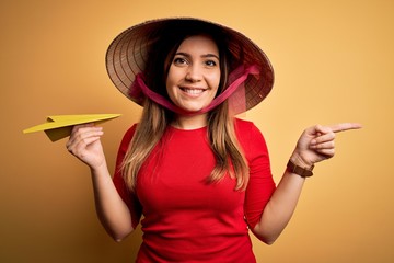 Poster - Tourist woman wearing traditional asian rice paddy straw hat holding paper plane for a trip very happy pointing with hand and finger to the side