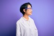 Young beautiful asian scientist girl wearing coat and glasses over purple background looking away to side with smile on face, natural expression. Laughing confident.