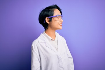 young beautiful asian scientist girl wearing coat and glasses over purple background looking away to