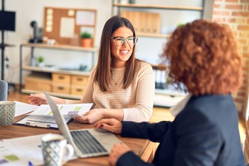 Two beautiful businesswomen smiling happy and confident. Sitting with smile on face working together using laptop at the office