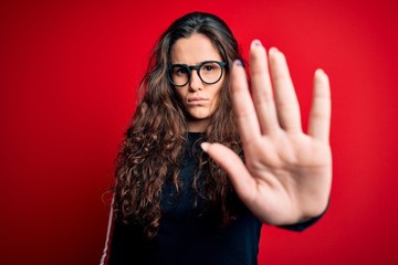 Poster - Young beautiful woman with curly hair wearing sweater and glasses over red background doing stop sing with palm of the hand. Warning expression with negative and serious gesture on the face.