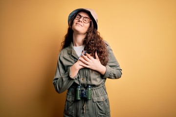 Poster - Young beautiful tourist woman on vacation wearing explorer hat and binoculars smiling with hands on chest with closed eyes and grateful gesture on face. Health concept.