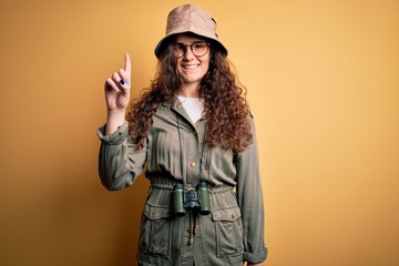 Poster - Young beautiful tourist woman on vacation wearing explorer hat and binoculars showing and pointing up with finger number one while smiling confident and happy.