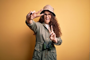 Poster - Young beautiful tourist woman on vacation wearing explorer hat and binoculars smiling looking to the camera showing fingers doing victory sign. Number two.