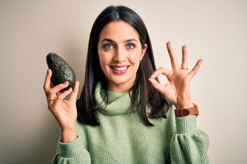 Young woman with blue eyes holding healthy avocado over isolated white background doing ok sign with fingers, excellent symbol