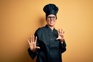 Poster - Young beautiful brunette chef woman wearing cooker uniform and hat over yellow background disgusted expression, displeased and fearful doing disgust face because aversion reaction. With hands raised