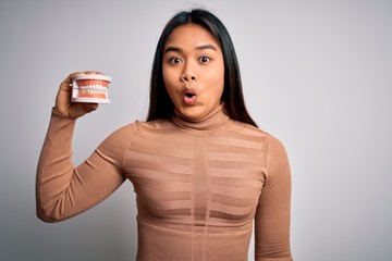 Young asian girl asking for dental care holding plastic denture teeth over white background scared in shock with a surprise face, afraid and excited with fear expression