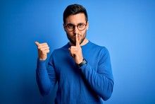 Young Handsome Man With Beard Wearing Casual Sweater And Glasses Over Blue Background Asking To Be Quiet With Finger On Lips Pointing With Hand To The Side. Silence And Secret Concept.