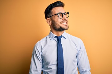 Young handsome businessman wearing tie and glasses standing over yellow background looking away to side with smile on face, natural expression. Laughing confident.