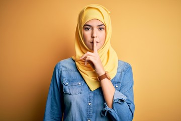 Sticker - Young beautiful girl wearing muslim hijab standing over isolated yellow background asking to be quiet with finger on lips. Silence and secret concept.