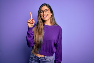 Wall Mural - Young beautiful smart woman wearing glasses over purple isolated background pointing finger up with successful idea. Exited and happy. Number one.