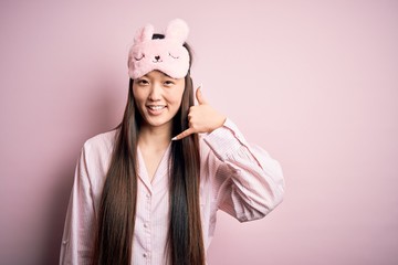 Sticker - Young asian woman wearing pajama and sleep mask over pink isolated background smiling doing phone gesture with hand and fingers like talking on the telephone. Communicating concepts.