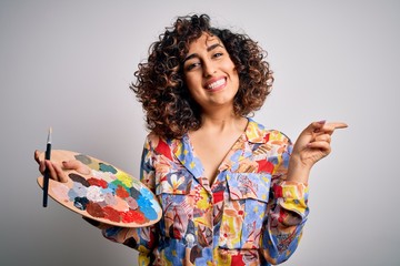 Sticker - Young beautiful curly arab artist woman painting using brush and palette with colors very happy pointing with hand and finger to the side