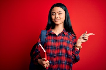 Wall Mural - Young beautiful chinese student woman holding book standing over isolated pink background very happy pointing with hand and finger to the side