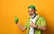 Grandpa In Green Cap And Shirt, White T-shirt, Bracelet. Holding Two Dumbbells And Pointing At Them, Posing On Orange Background