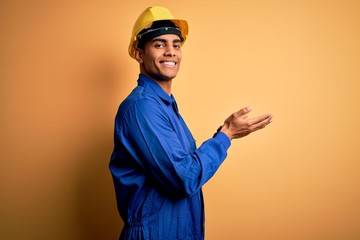 Wall Mural - Young handsome african american worker man wearing blue uniform and security helmet pointing aside with hands open palms showing copy space, presenting advertisement smiling excited happy