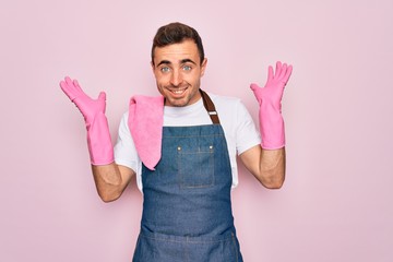 Wall Mural - Young cleaner man with blue eyes cleaning wearing apron and gloves over pink background celebrating crazy and amazed for success with arms raised and open eyes screaming excited. Winner concept