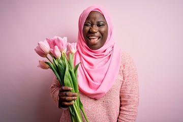 Sticker - Young african american plus size woman wearing muslim hijab holding bouquet of pink tulips winking looking at the camera with sexy expression, cheerful and happy face.